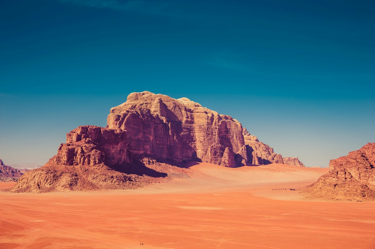 A large sandstone mountain rising from red desert sand under a deep blue sky in Wadi Rum